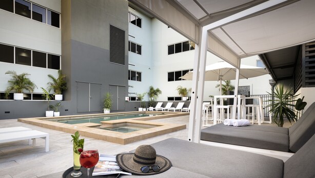 A modern indoor pool area with lounge chairs, a pool table, and a seating area with a hat and sunglasses on a tray. - Grand Hotel and Apartments Townsville (South Townsville)