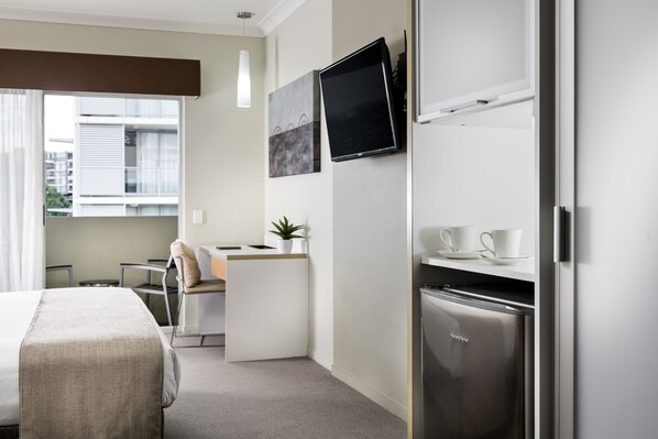 A hotel room with a bed, a desk, a TV, a refrigerator, and a window with a view of buildings.