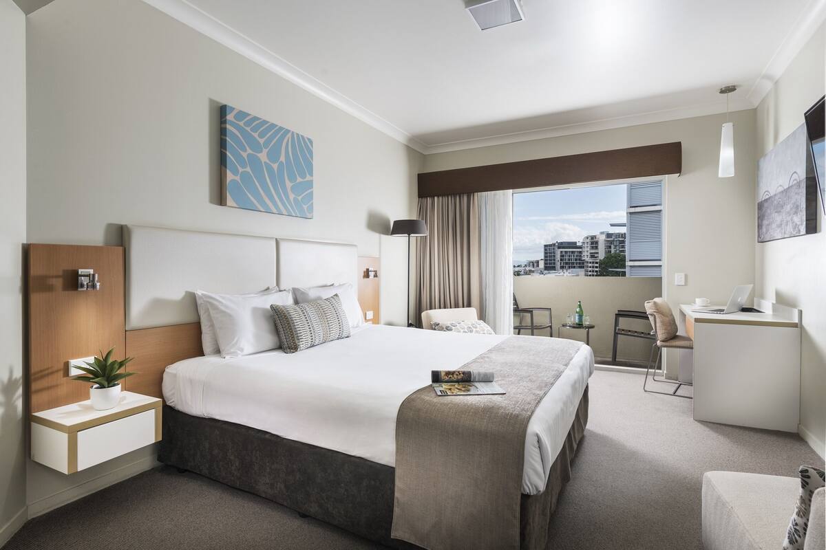 Ein modernes Hotelzimmer mit einem großen Bett, einem Schreibtisch, einem Sessel, einem kleinen Tisch mit Pflanze und Blick auf die Stadt durch das Fenster.