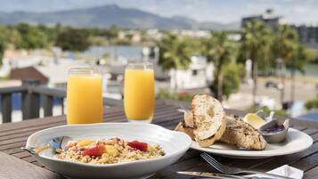 A breakfast table with a bowl of cereal, toast, and a glass of orange juice, overlooking a marina with boats and mountains in the background.