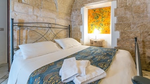 A bedroom with a stone wall, a bed with white linens, a blue patterned bedspread, and a painting on the wall.
