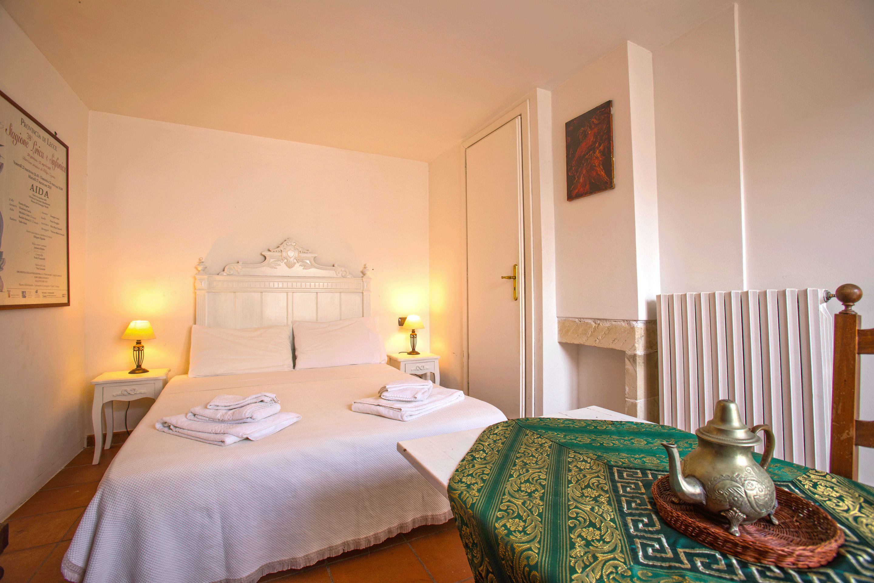 Una cama bien hecha con sábanas blancas, dos lámparas de noche, un plaid verde estampado y un jarro metálico decorativo sobre una bandeja tejida.