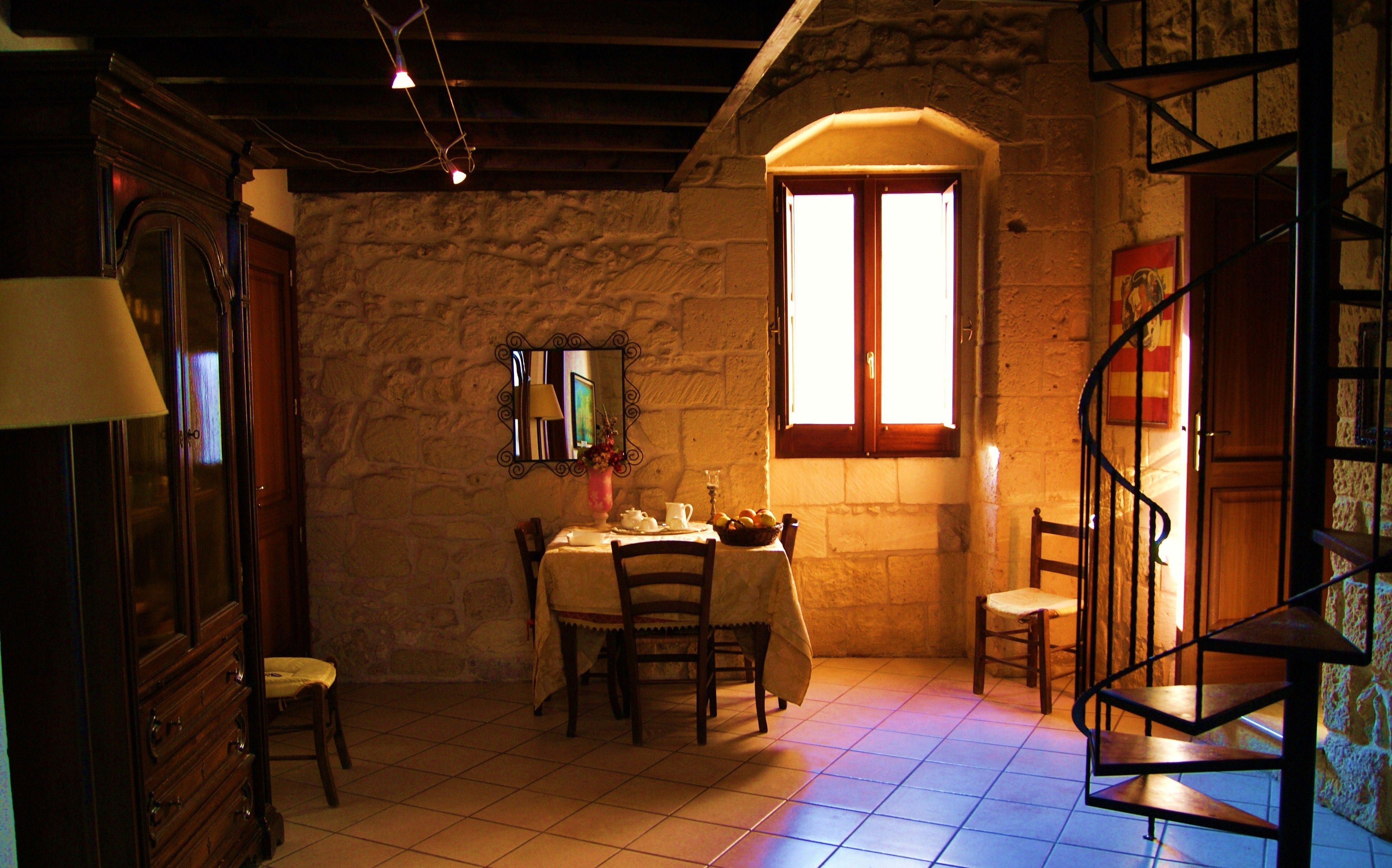 A dining area with a stone wall, a window with wooden shutters, a table set with a vase of flowers, and a chair.
