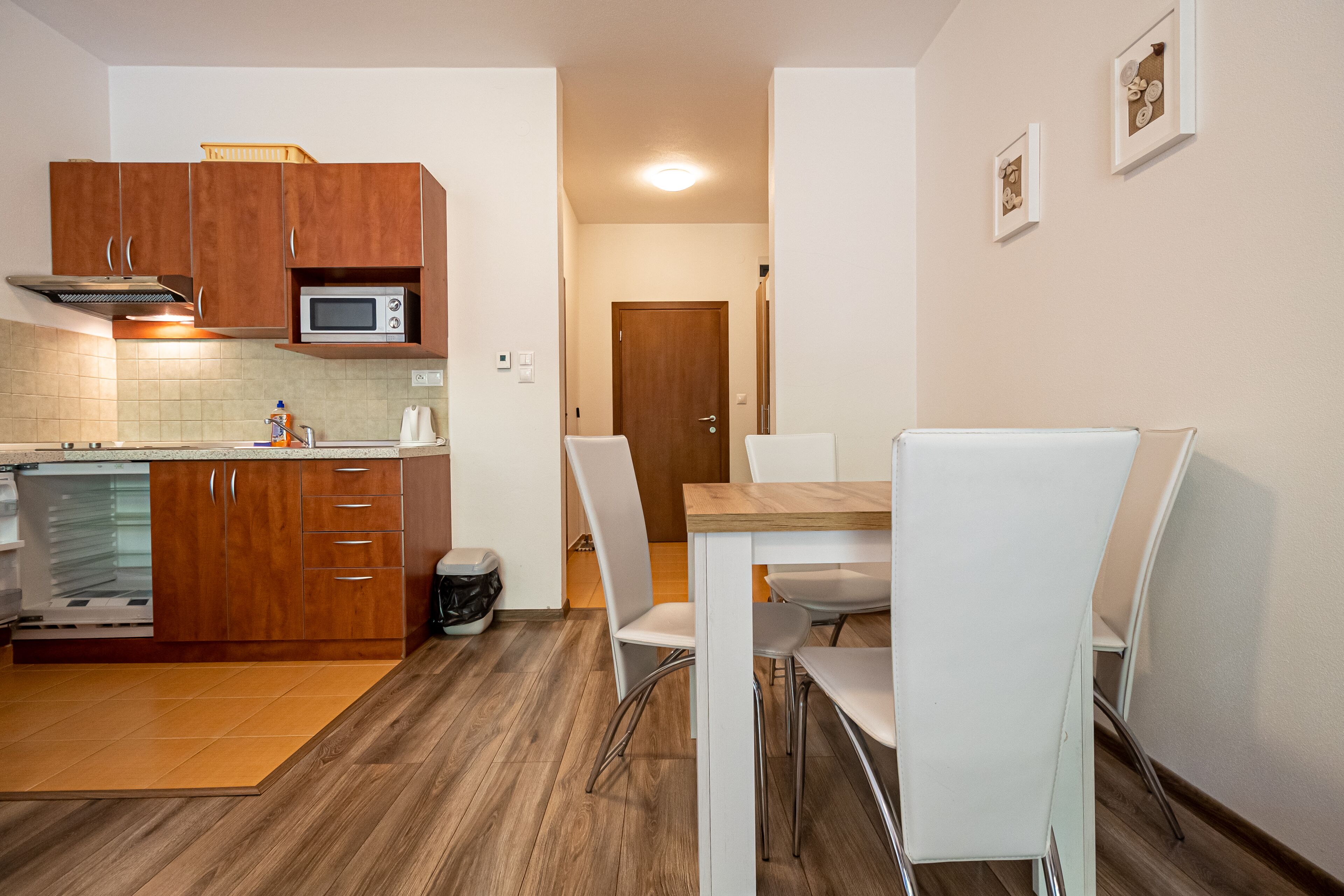 Cocina compacta con armarios de madera, microondas y zona de comedor con sillas blancas y una mesa.