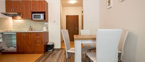 Cocina compacta con armarios de madera, microondas y zona de comedor con sillas blancas y una mesa.