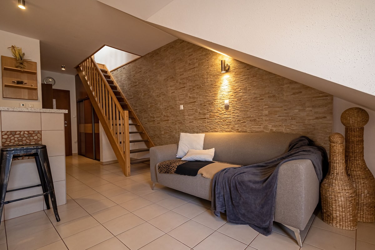 A modern loft with a sloped ceiling, a staircase, a sofa, and a kitchen area.