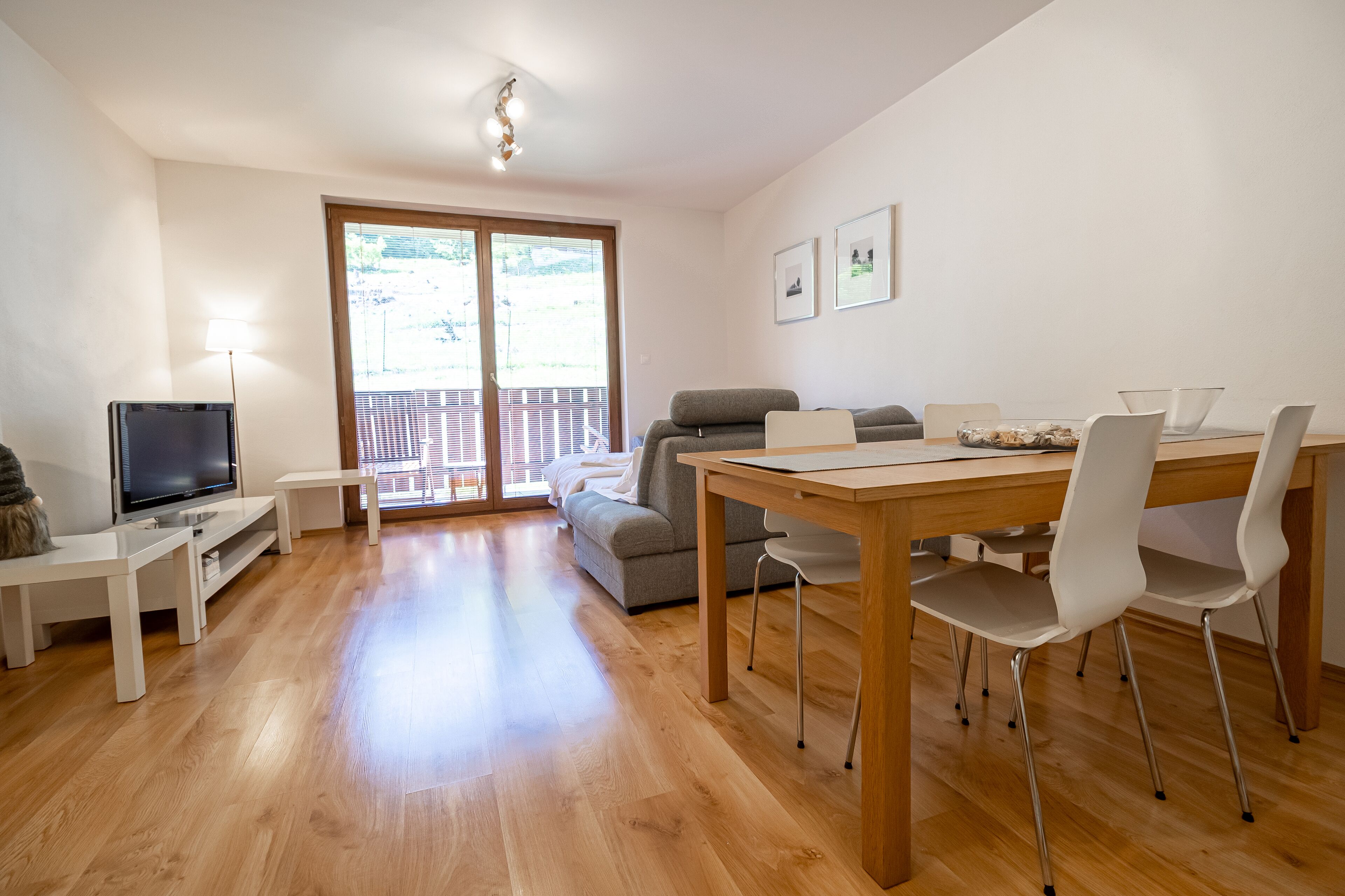 Una sala moderna con piso de madera, televisor, sofá, mesa de comedor con sillas y balcón con vistas.