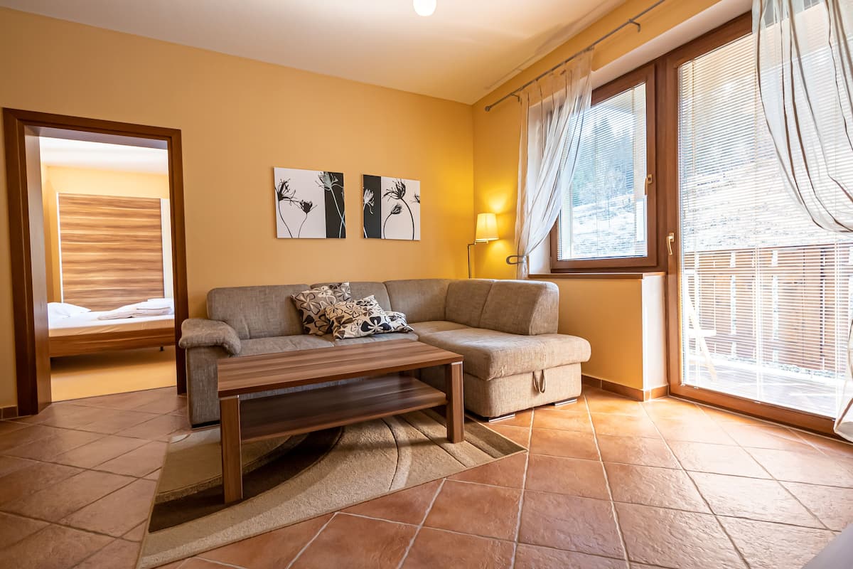 A living room with a sofa, a coffee table, and a lamp. There are two paintings on the wall and a window with curtains.