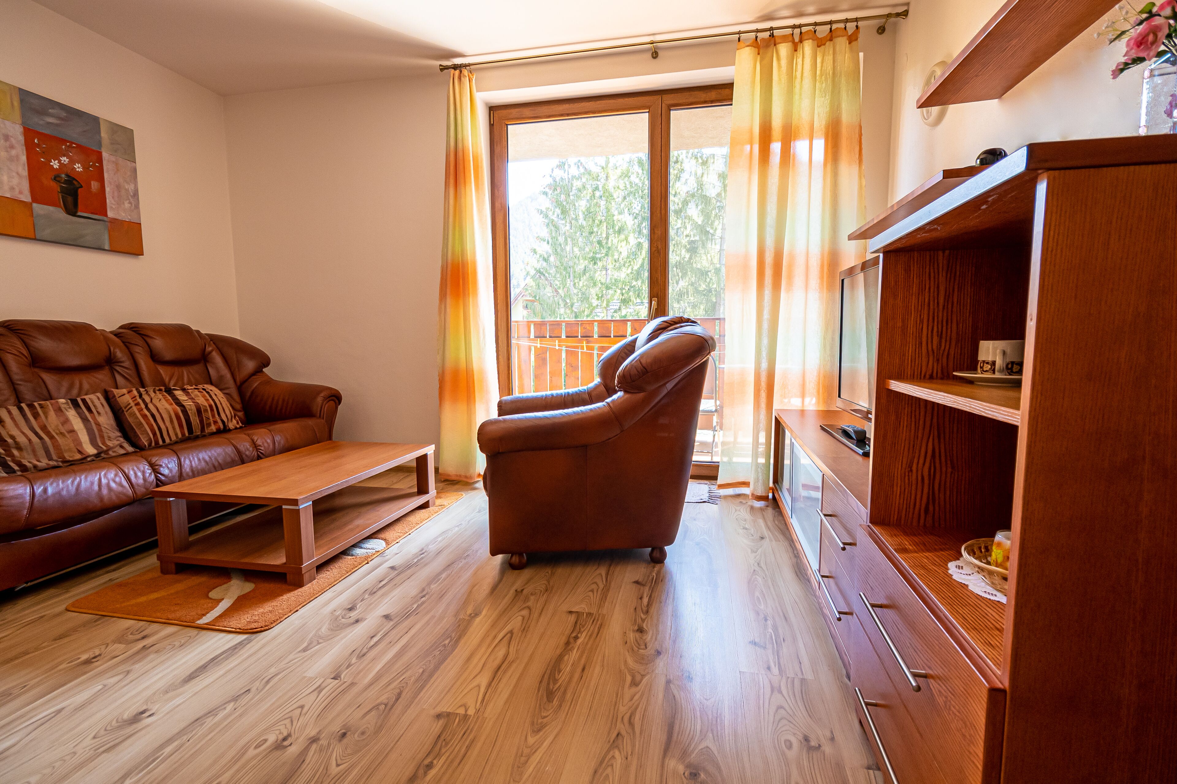 A living room with a leather sofa, a wooden coffee table, and a brown armchair. There is a wooden cabinet with drawers and a television on top. A window with curtains is visible.