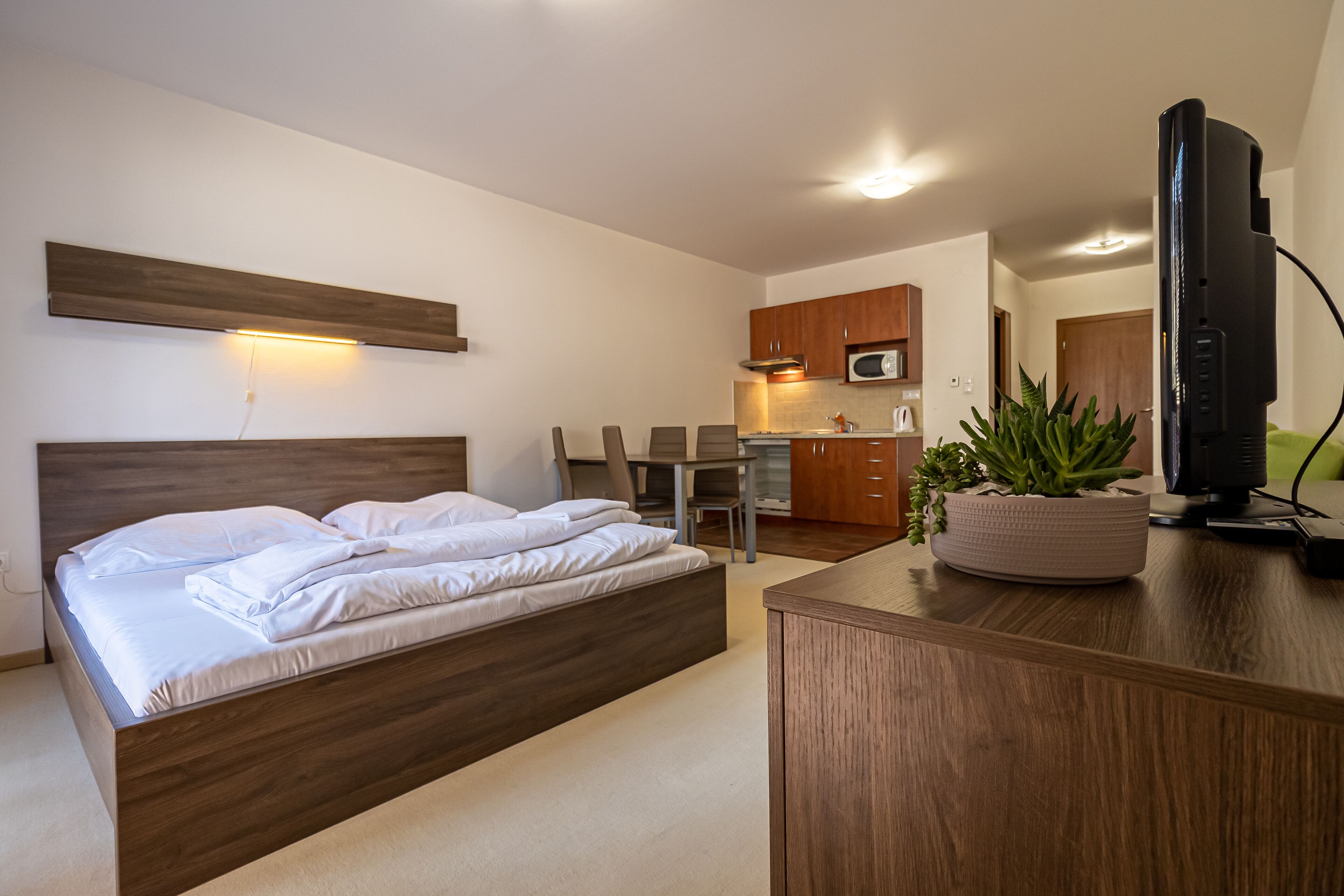 Un dormitorio moderno con una cama de madera, una pequeña mesa de comedor y una zona de cocina con armarios de madera y un microondas.