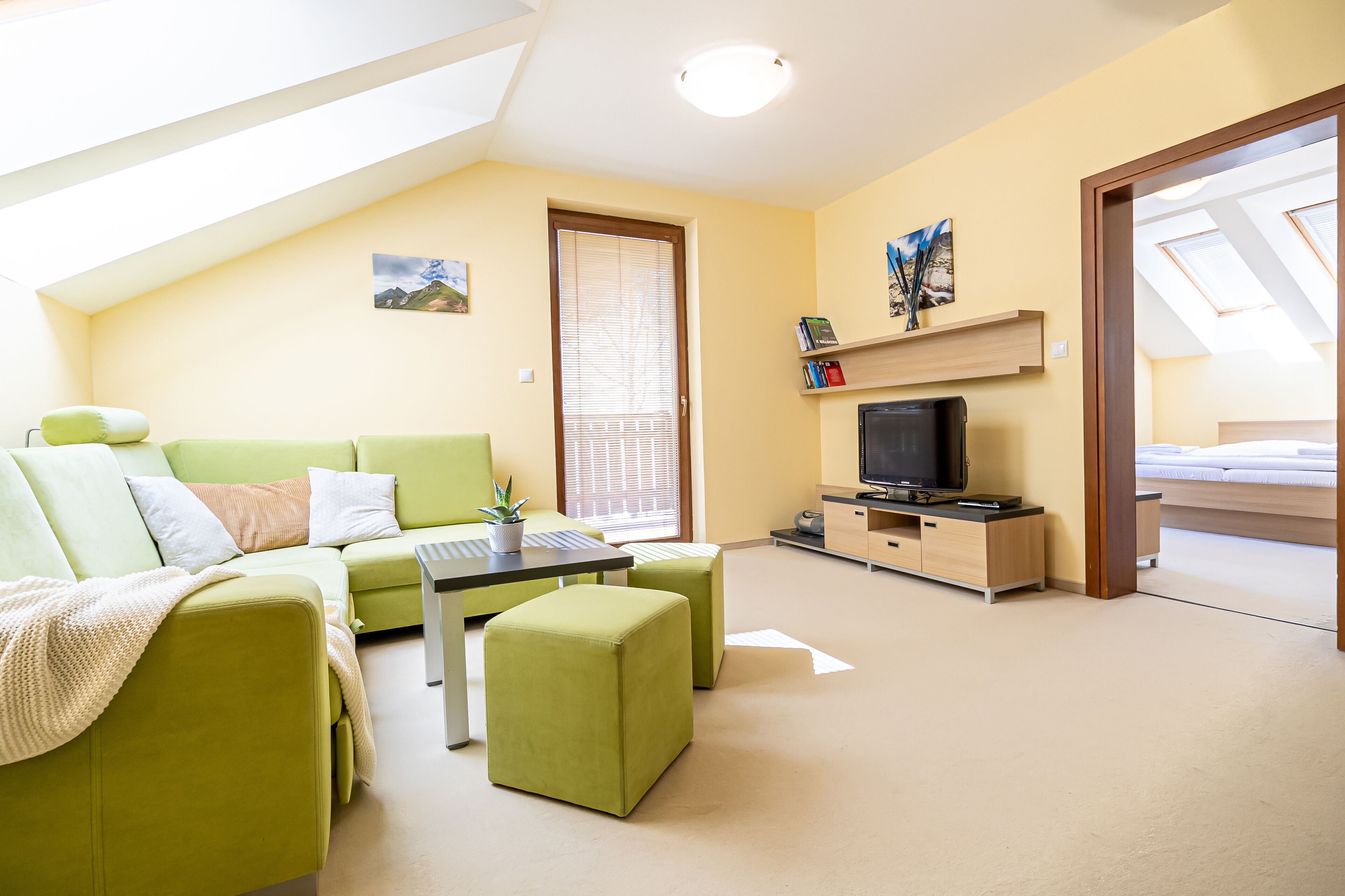 Ein modernes, helles Zimmer mit einem grünen Sofa, einem kleinen Tisch, einem Fernseher und einem Bett im Hintergrund.