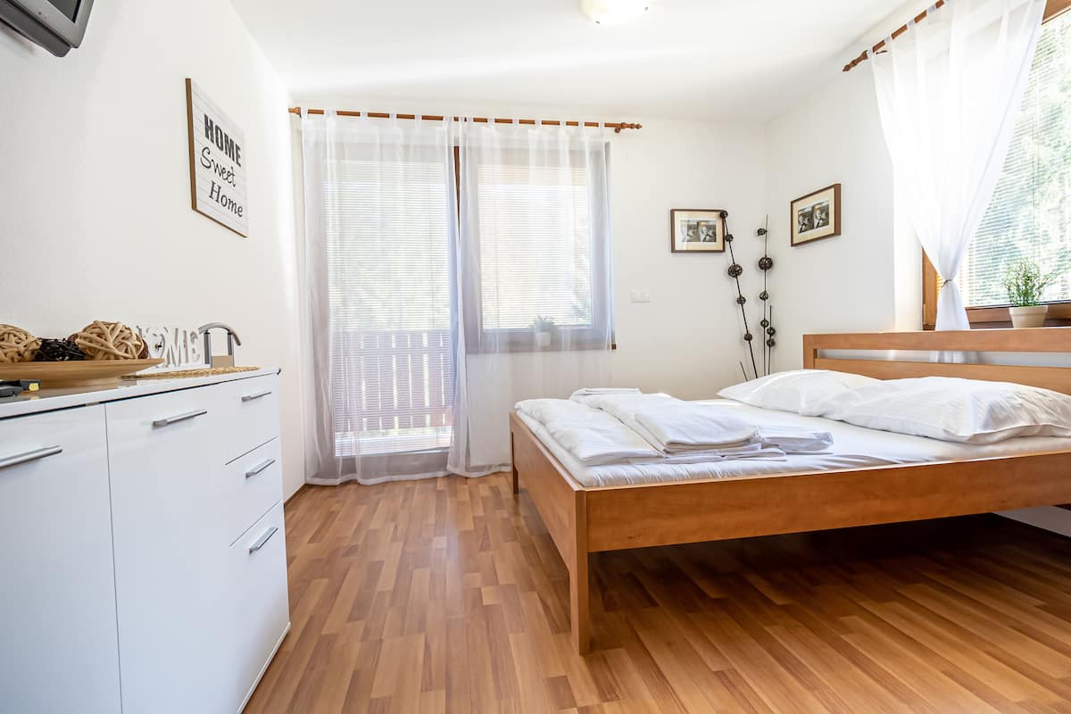 A bedroom with a wooden bed, white curtains, a small desk with a chair, and a window with blinds.