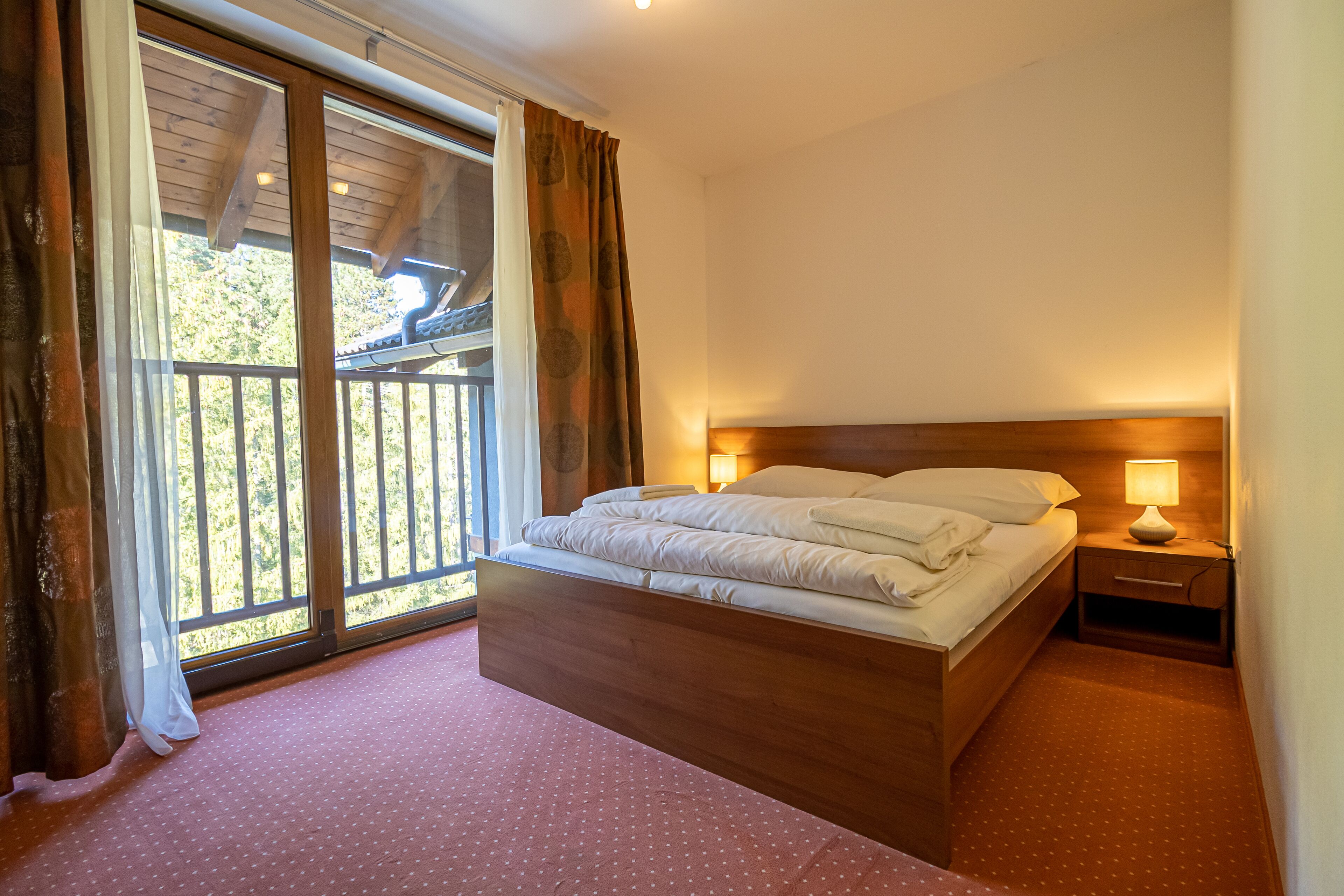 A bedroom with a wooden bed, a nightstand with a lamp, and a large window with curtains.