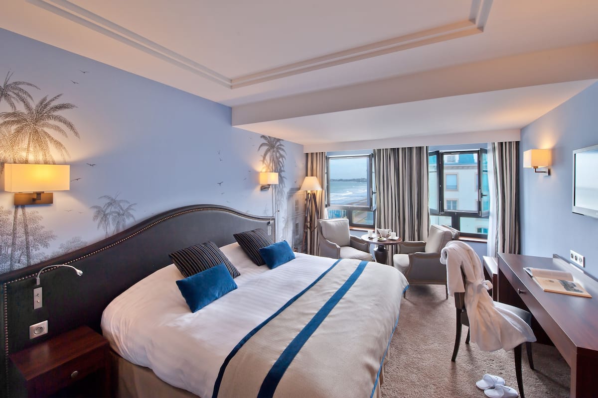 A hotel room with a large bed, a desk, a chair, and a view of the sea through the window.