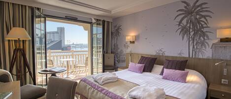 Une chambre d’hôtel avec un grand lit, un bureau, une chaise, un balcon offrant une vue sur la ville et des œuvres d’art murales représentant des palmiers.