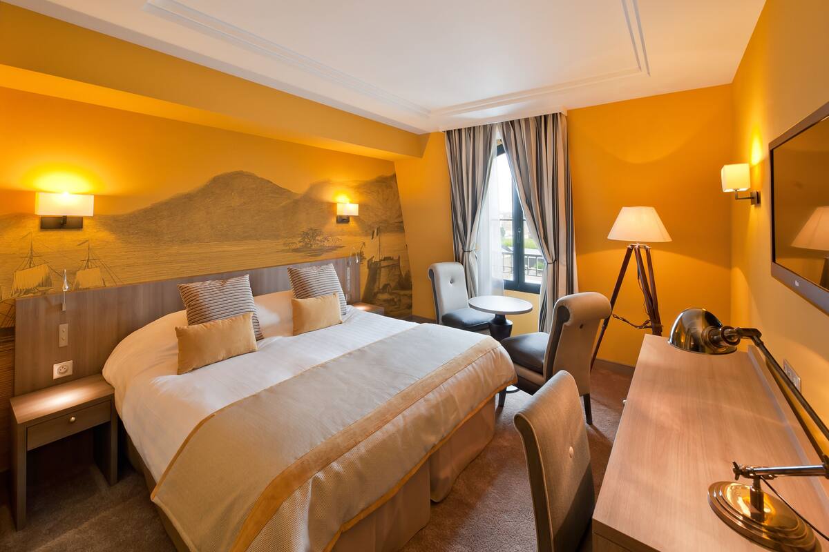 A hotel room with a large bed, a desk, a chair, a small table, and a lamp.