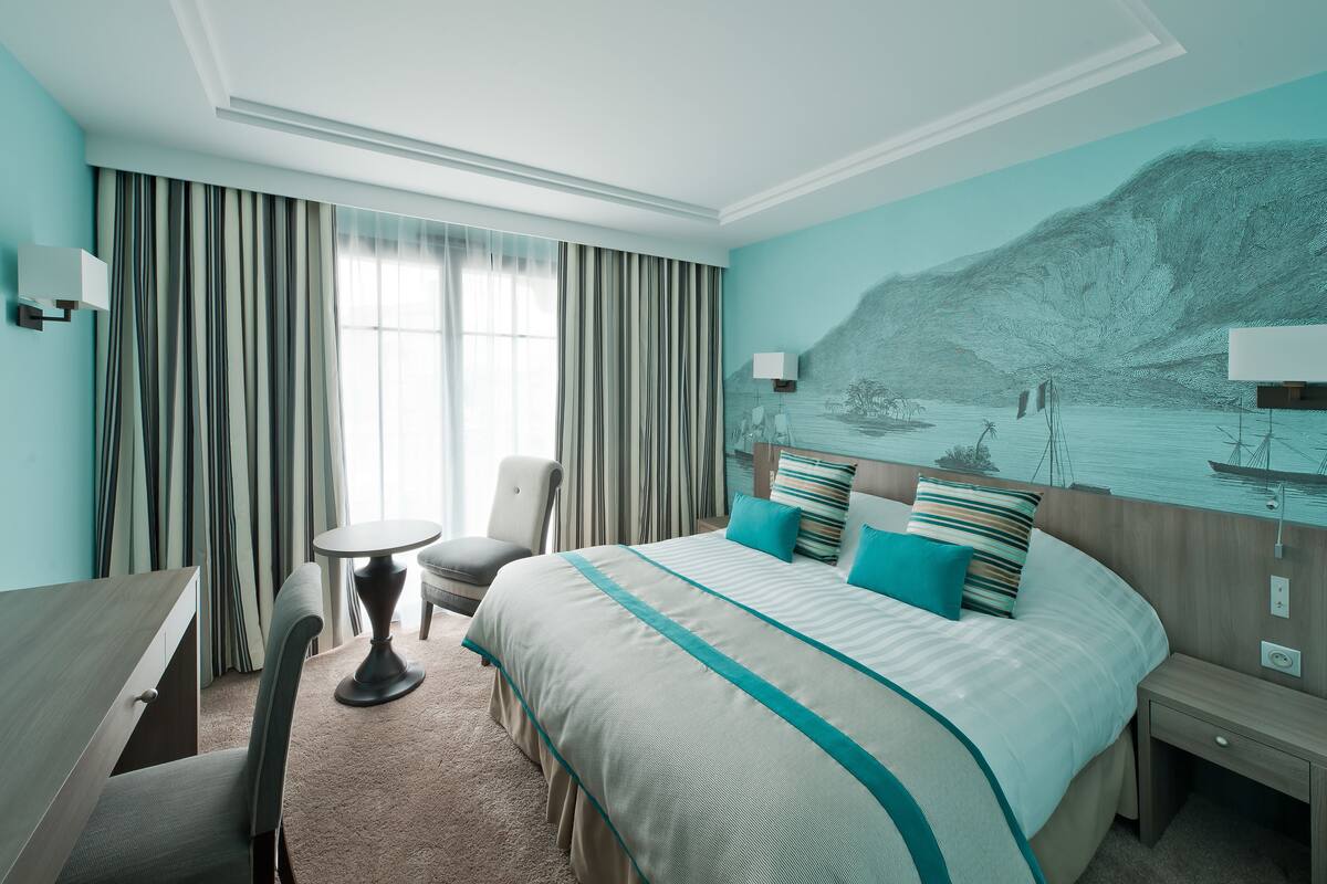 A hotel room with a large bed, a desk, a chair, and a view of a mountainous landscape through the window.
