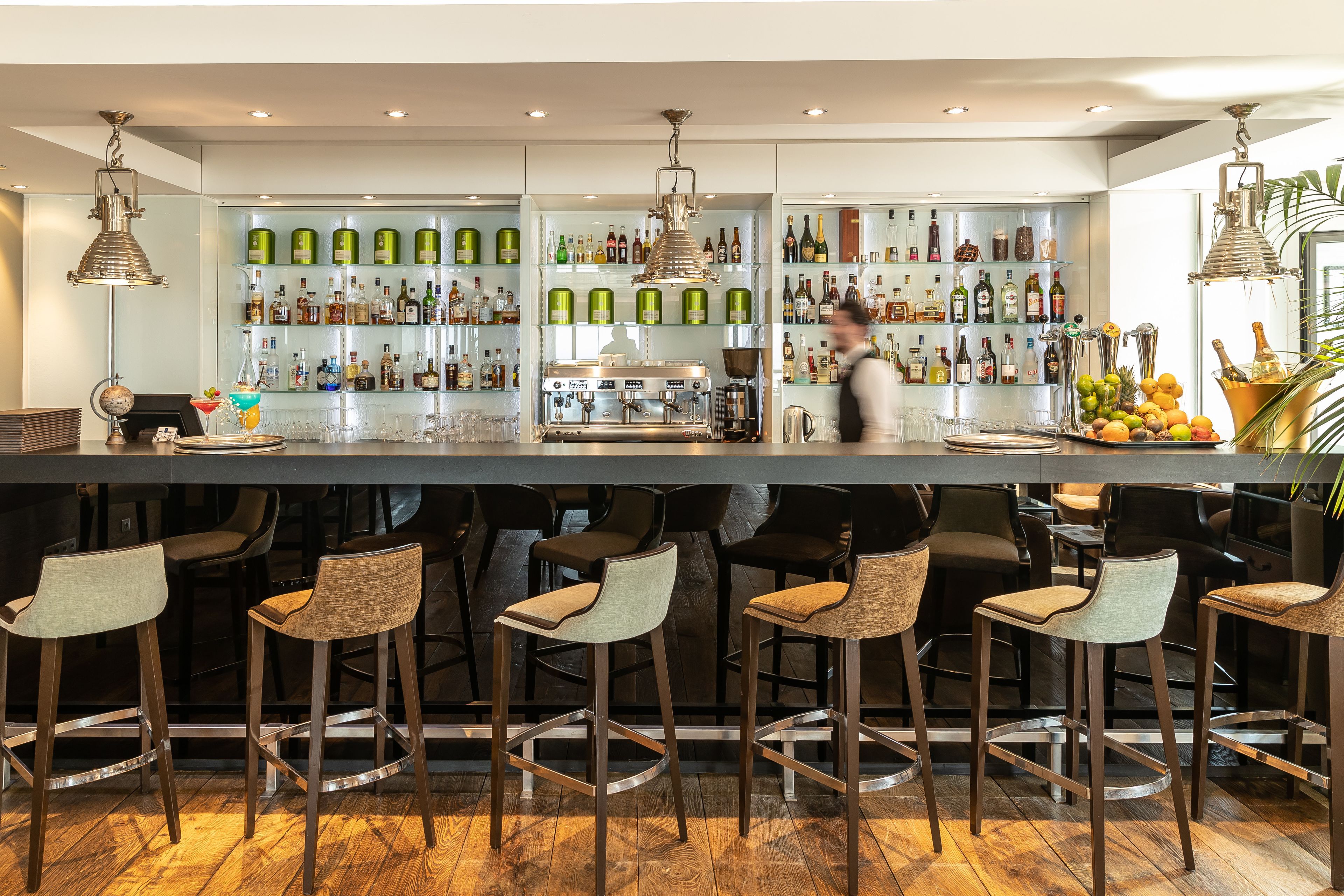 A bar with a polished wooden floor, multiple stools, and a well-stocked shelf with various bottles and glasses.