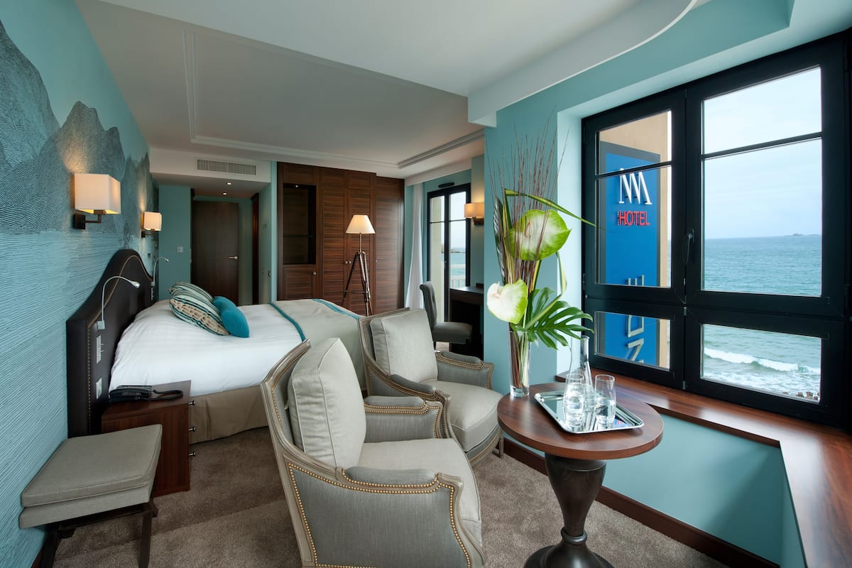 A hotel room with a bed, two armchairs, a small table with a vase, and a view of the sea through large windows.