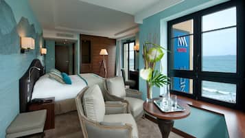 A hotel room with a bed, two armchairs, a small table with a vase, and a view of the sea through large windows.