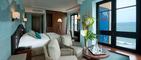 Habitación de hotel con una cama, dos sillones, una mesita con un jarrón y vistas al mar a través de amplias ventanas.
