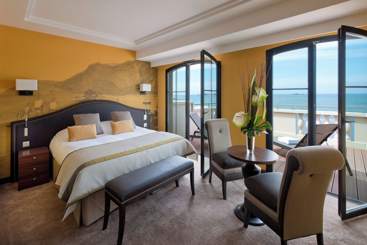 A hotel room with a bed, a bench, a chair, a table, and a view of the sea through large windows.