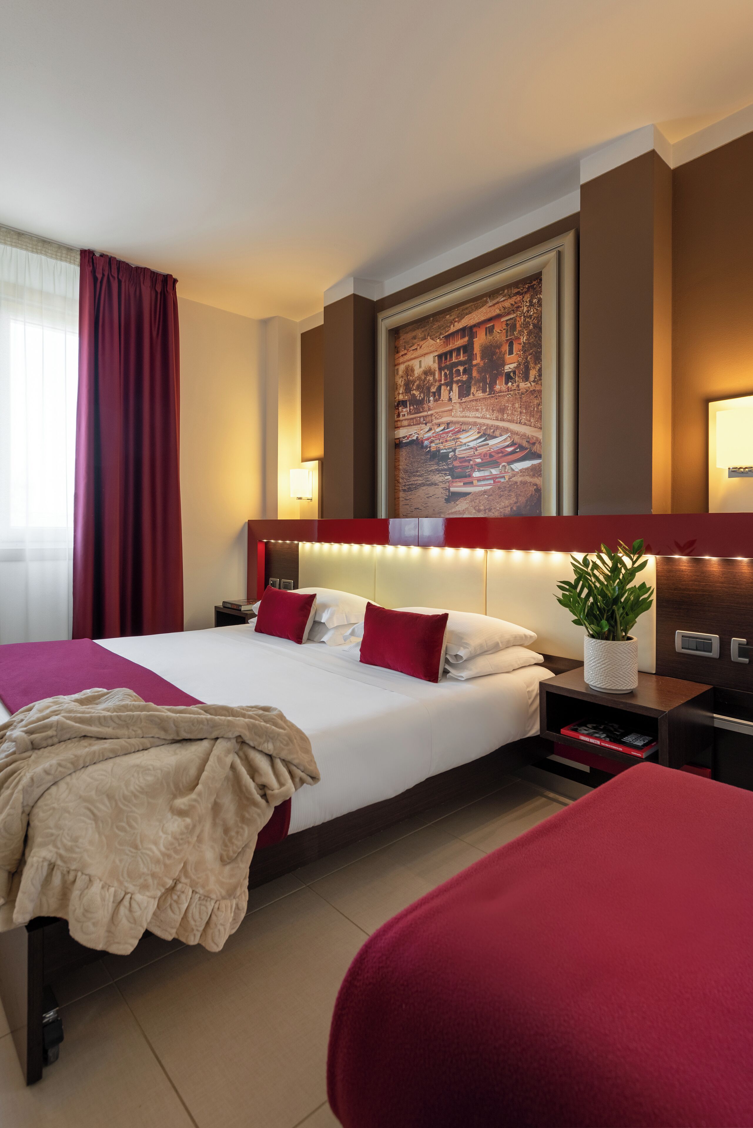 A hotel room with a large bed, red curtains, a headboard with integrated lighting, a nightstand with a lamp, a framed picture on the wall, and a potted plant.