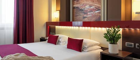 Habitación de hotel con una cama grande, cortinas rojas, cabecera con iluminación integrada, mesita de noche con lámpara, un cuadro en la pared y una planta en maceta.