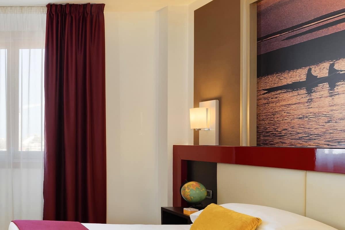 A hotel room with a large bed, a nightstand with a globe, a lamp, and a painting of a boat on water.