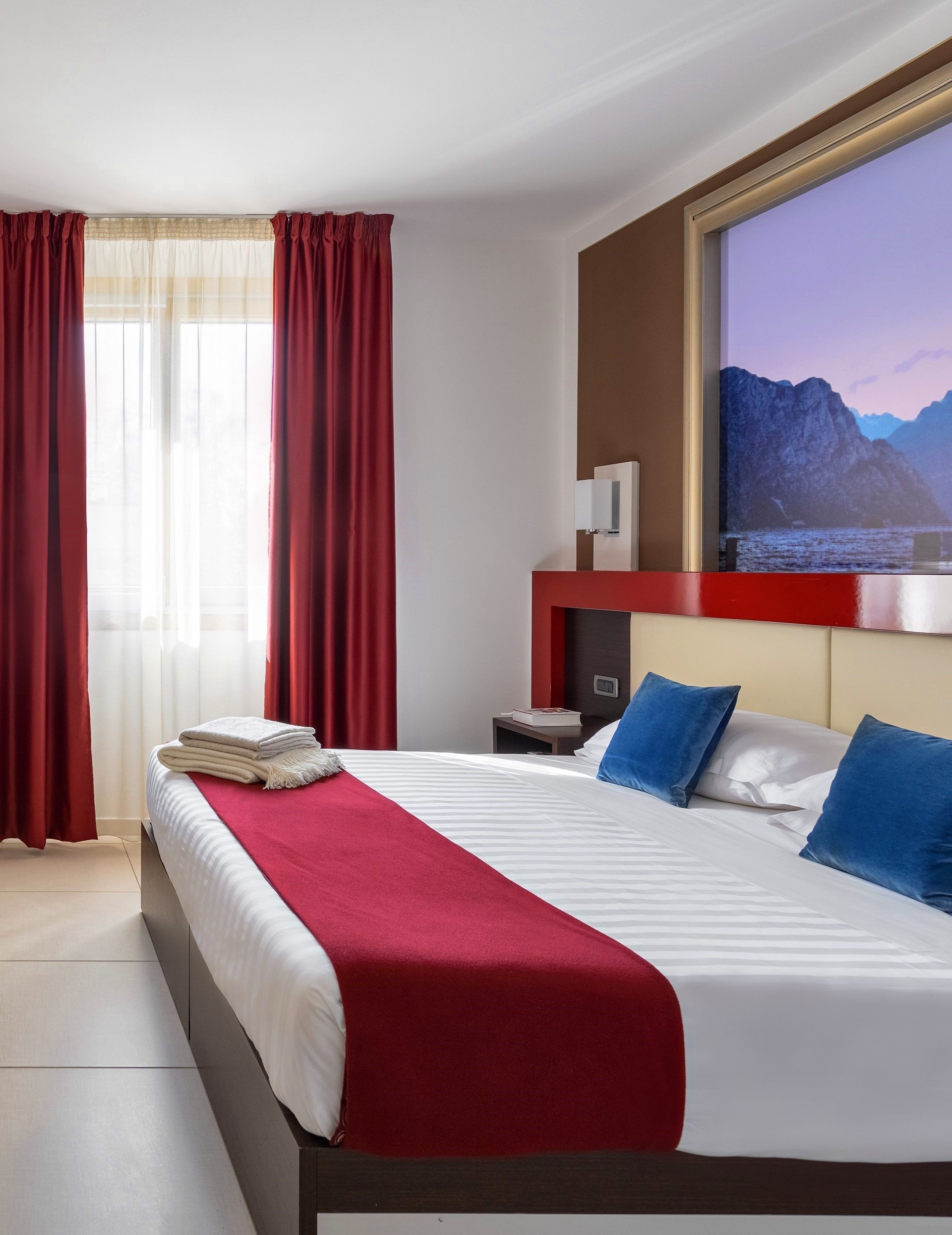 Une chambre d’hôtel avec un lit, des rideaux rouges, une grande fenêtre donnant sur la montagne, une table de chevet et un téléviseur fixé au mur.