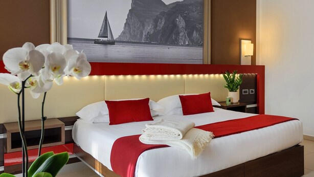 Habitación de hotel con una cama grande, ropa de cama roja y blanca, una mesita de noche con lámpara, una pequeña mesa con una revista y un cuadro grande enmarcado de un velero en la pared.