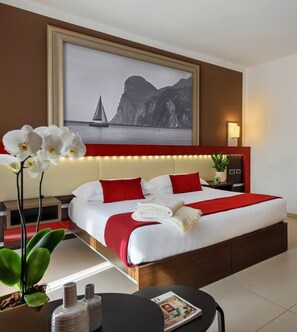 A hotel room with a large bed, red and white bedding, a nightstand with a lamp, a small table with a magazine, and a large framed picture of a sailboat on the wall. - Kairos Garda Hotel - Be Happy Hotels (Castelnuovo del Garda)