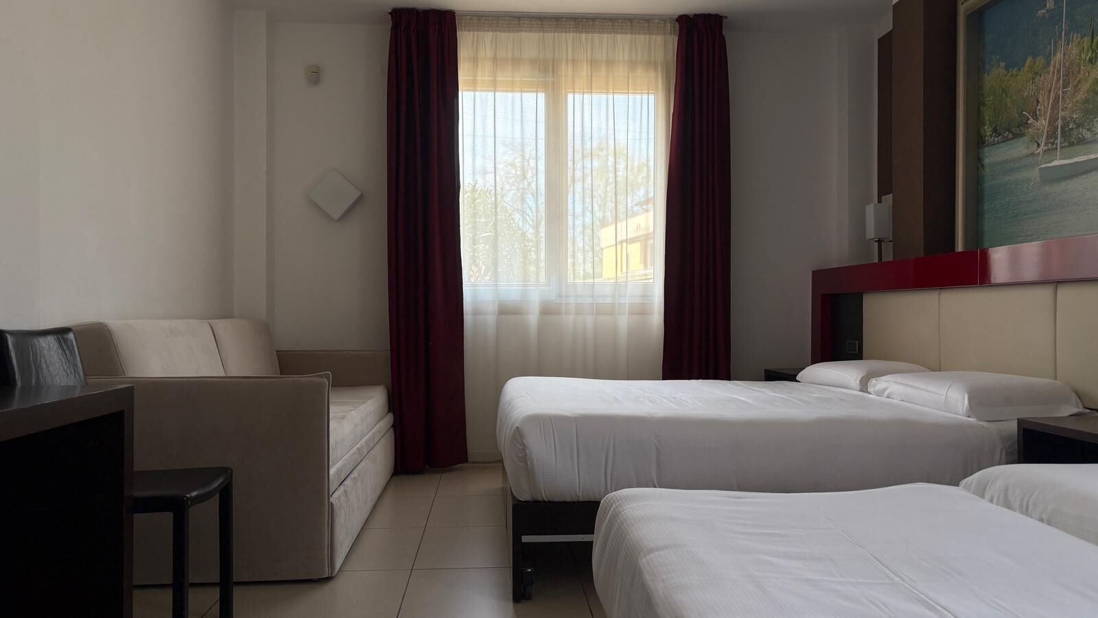 A hotel room with a sofa, two single beds, a desk, and a painting on the wall.
