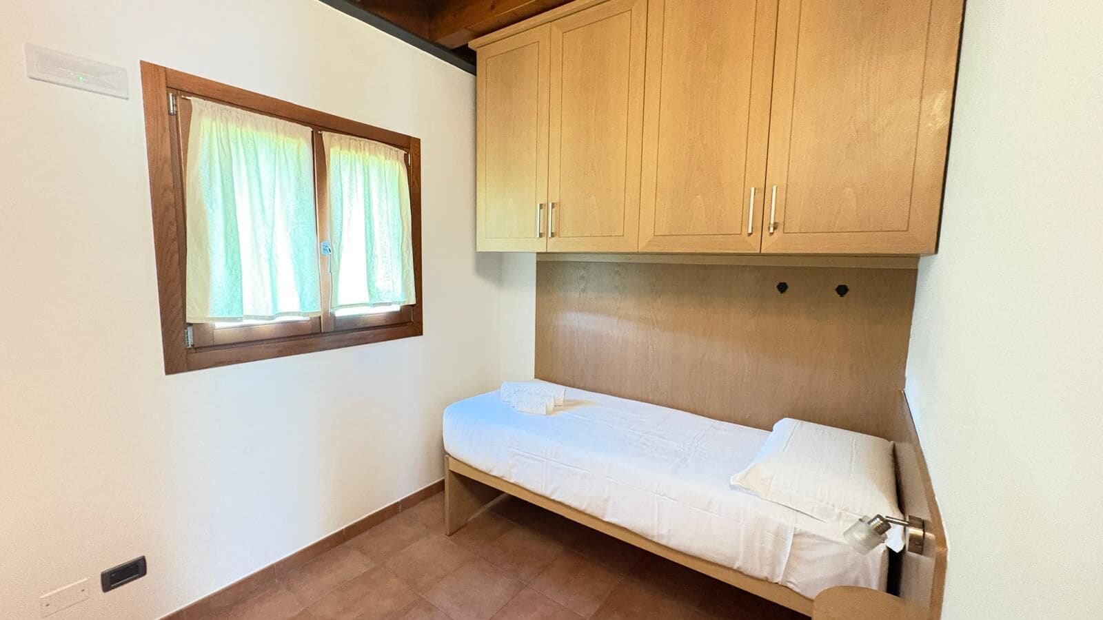 Una habitación pequeña con una cama individual, armarios de madera, una ventana con cortinas y un interruptor de luz en la pared.