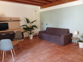 Una sala de estar con un sofá marrón, un televisor, una planta en maceta y una mesita de noche con una lámpara.