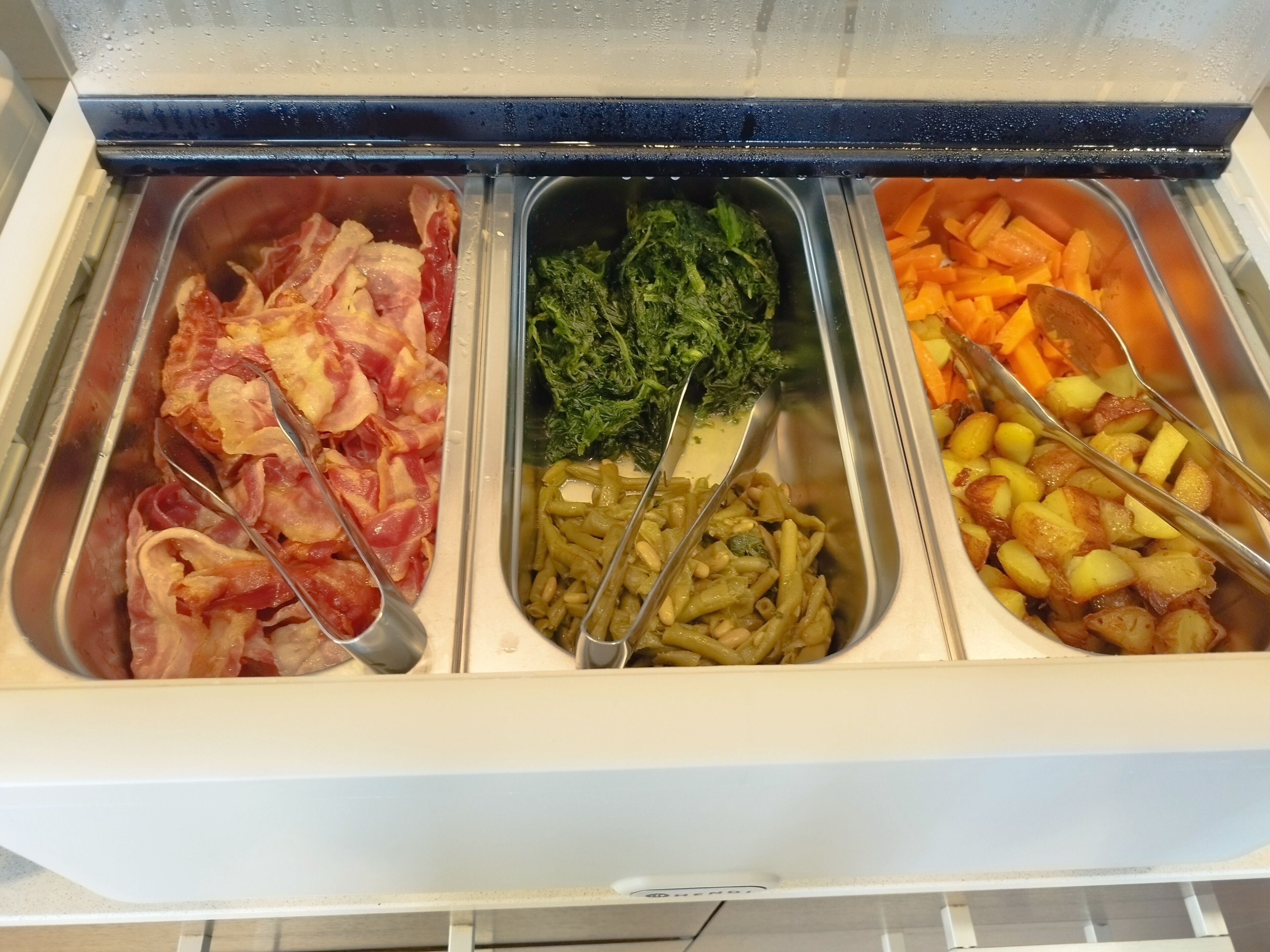 Un bufé con tres fuentes de comida: tocino, verduras y papas.