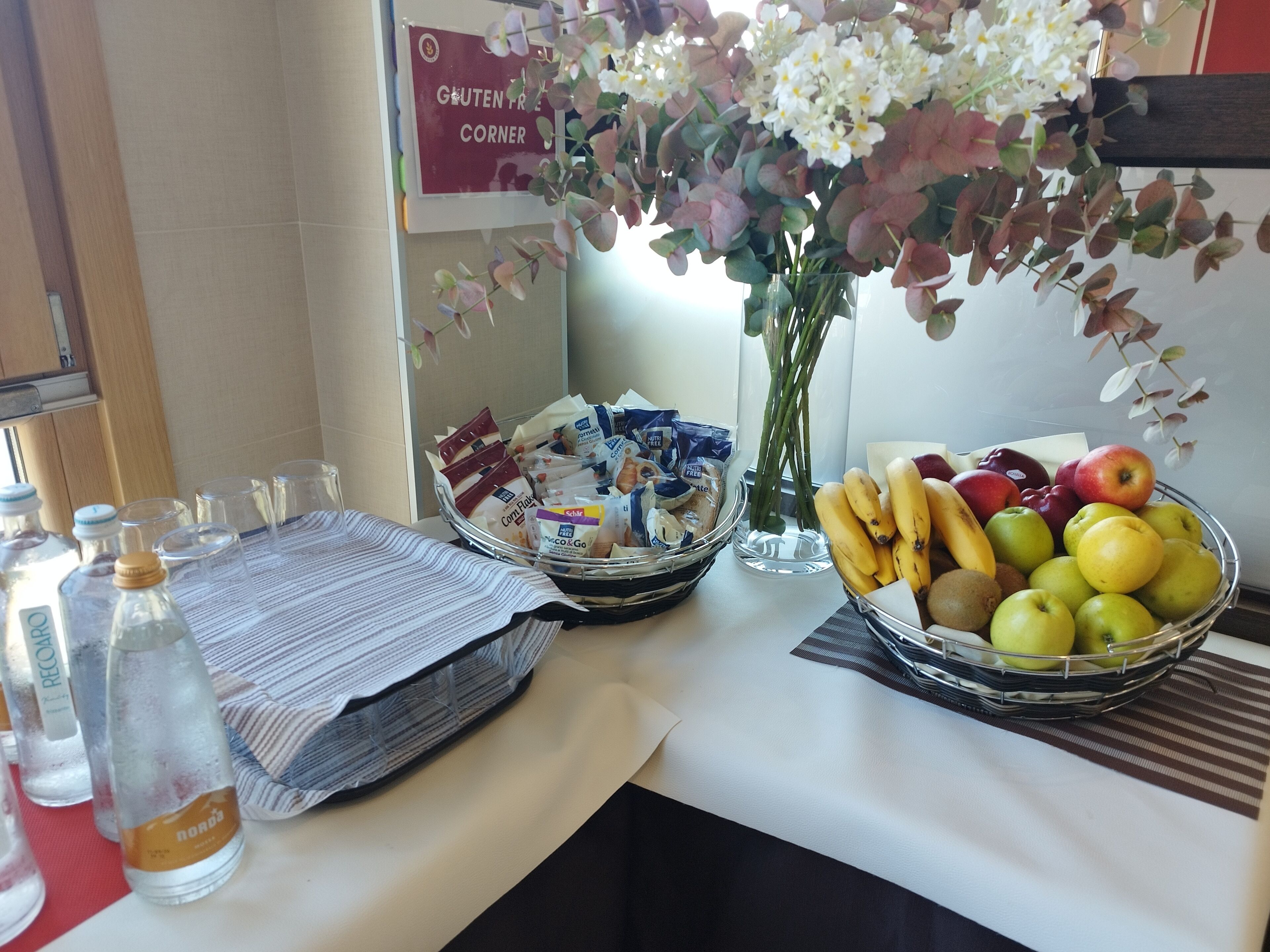 Ein Tisch mit verschiedenen Speisen, darunter Obst, verpackte Snacks, eine Vase mit Blumen und ein Schild, das auf einen glutenfreien Bereich hinweist.
