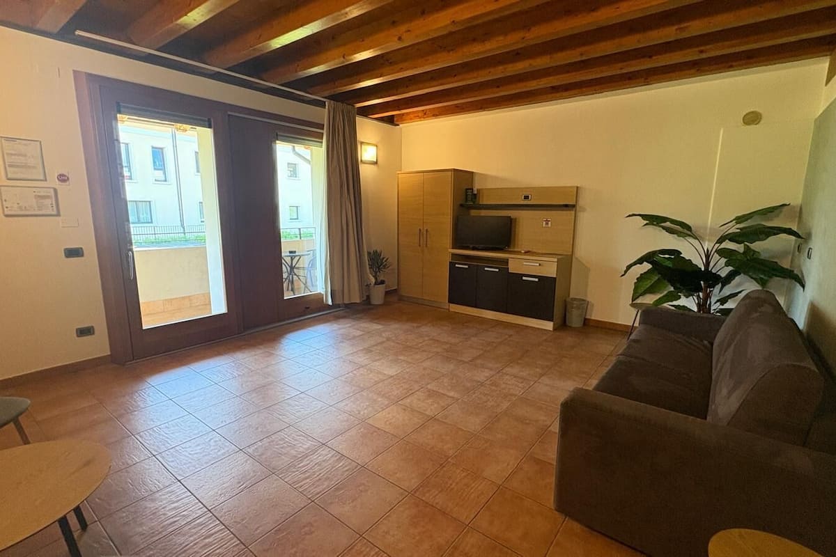 A living room with a sofa, a small table, a kitchen area, and a sliding glass door leading to an outdoor balcony.