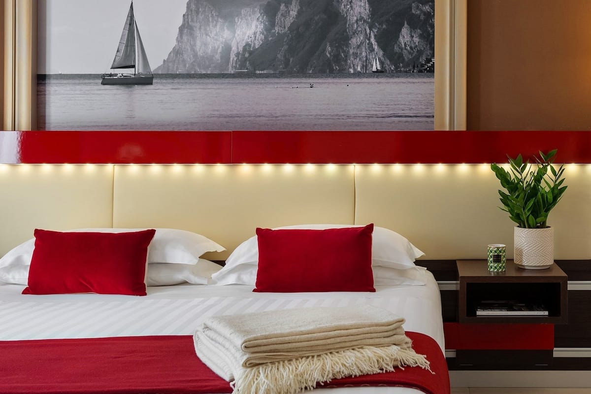 A hotel room with a bed, red and white bedding, a nightstand with a plant, and a large framed picture of a sailboat on a cliff.