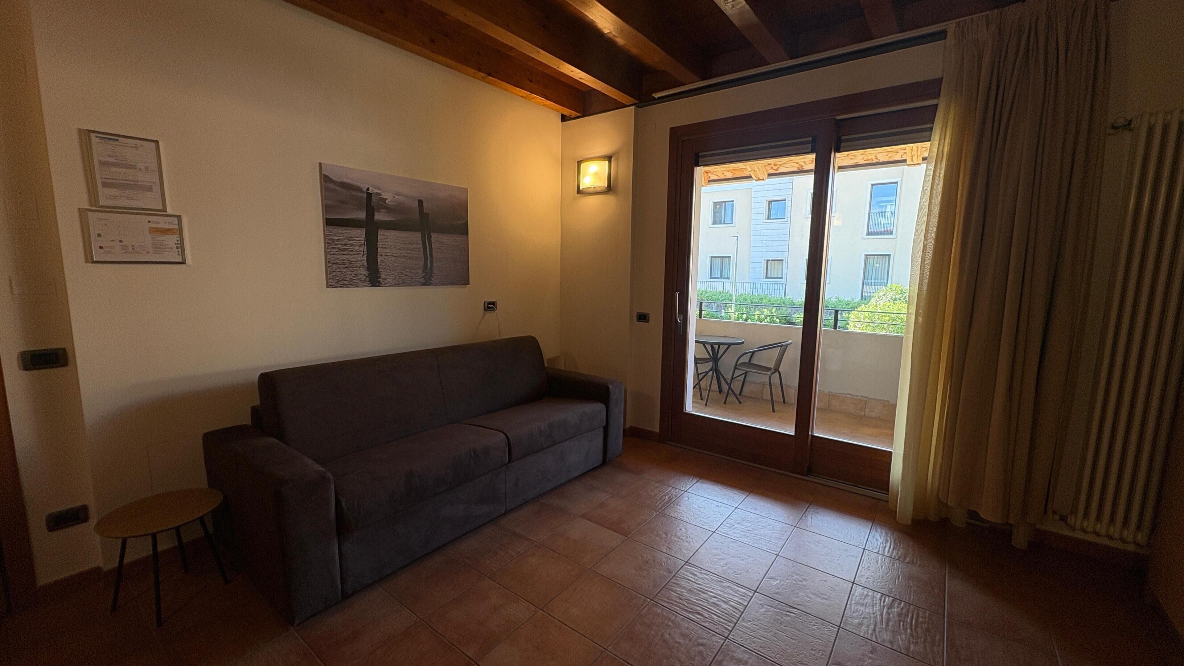 A room with a sofa, a small table, a sliding glass door leading to an outdoor area with a table and chairs, and a framed picture on the wall.