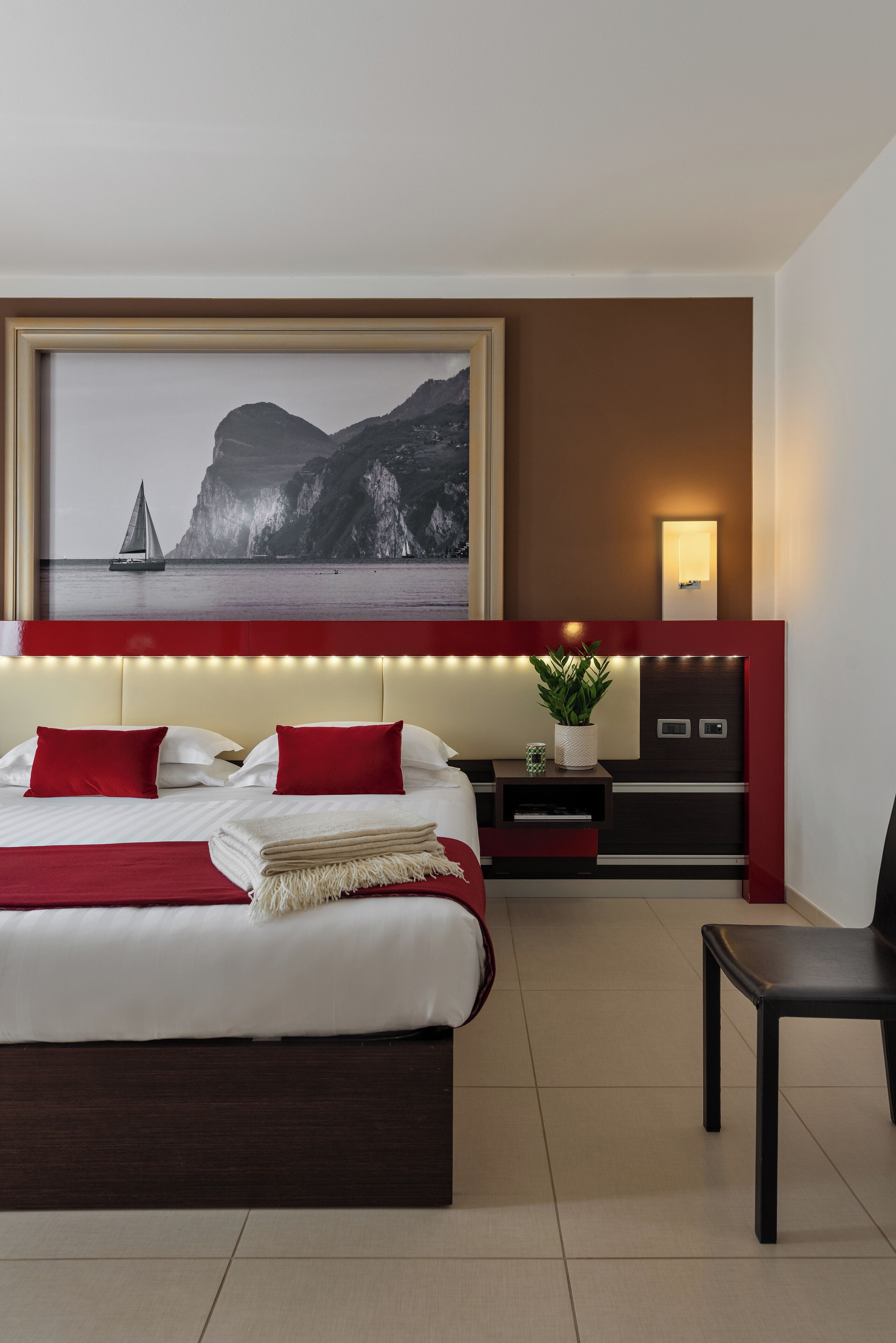 Habitación de hotel con una cama grande, almohadas rojas, una mesita de noche con una planta, una lámpara fijada a la pared y un cuadro enmarcado con un velero y acantilados.