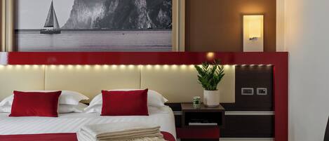 Habitación de hotel con una cama grande, almohadas rojas, una mesita de noche con una planta, una lámpara fijada a la pared y un cuadro enmarcado con un velero y acantilados.