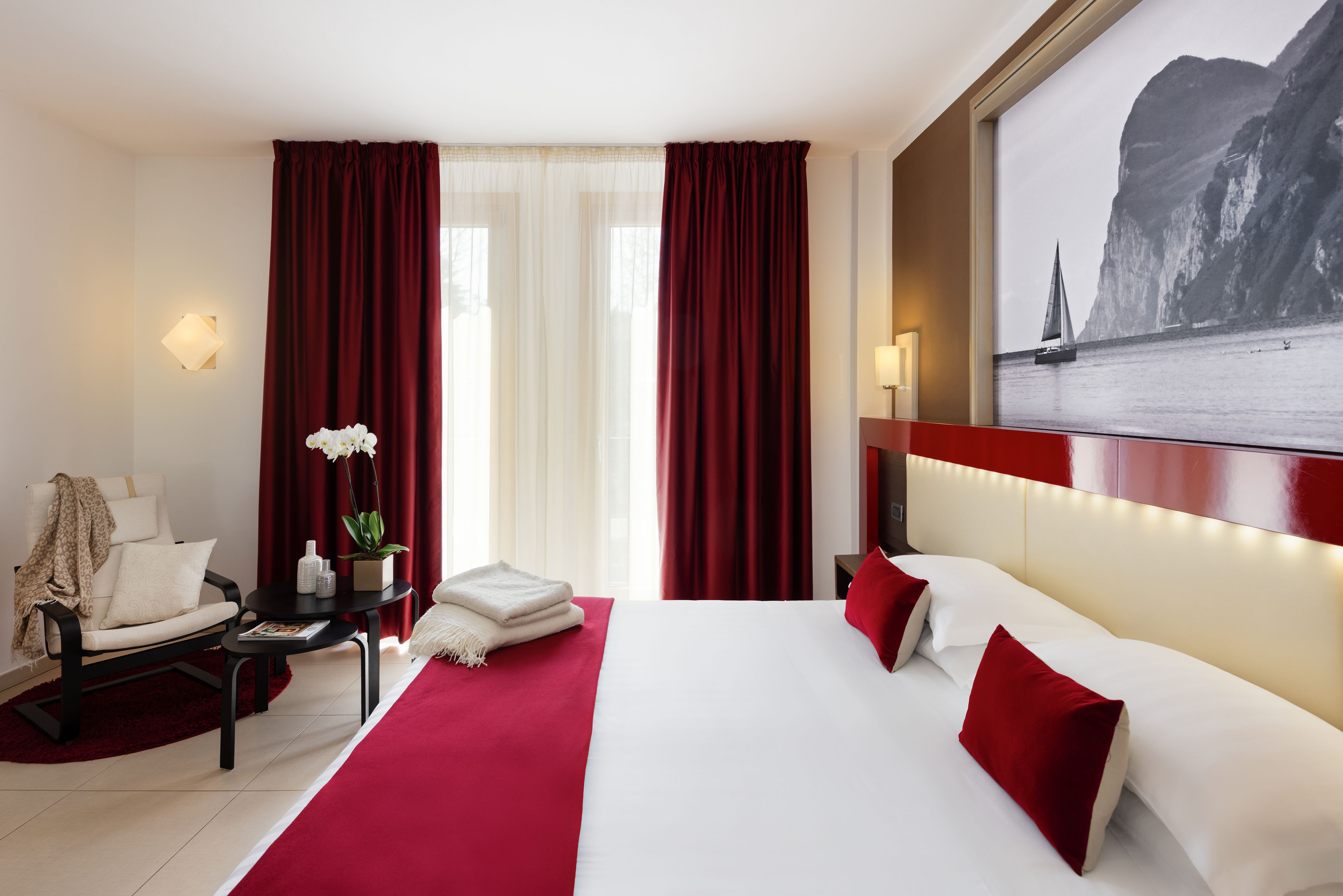 A hotel room with a large bed, red curtains, a desk with a chair, a small table with a plant, and a wall-mounted lamp.