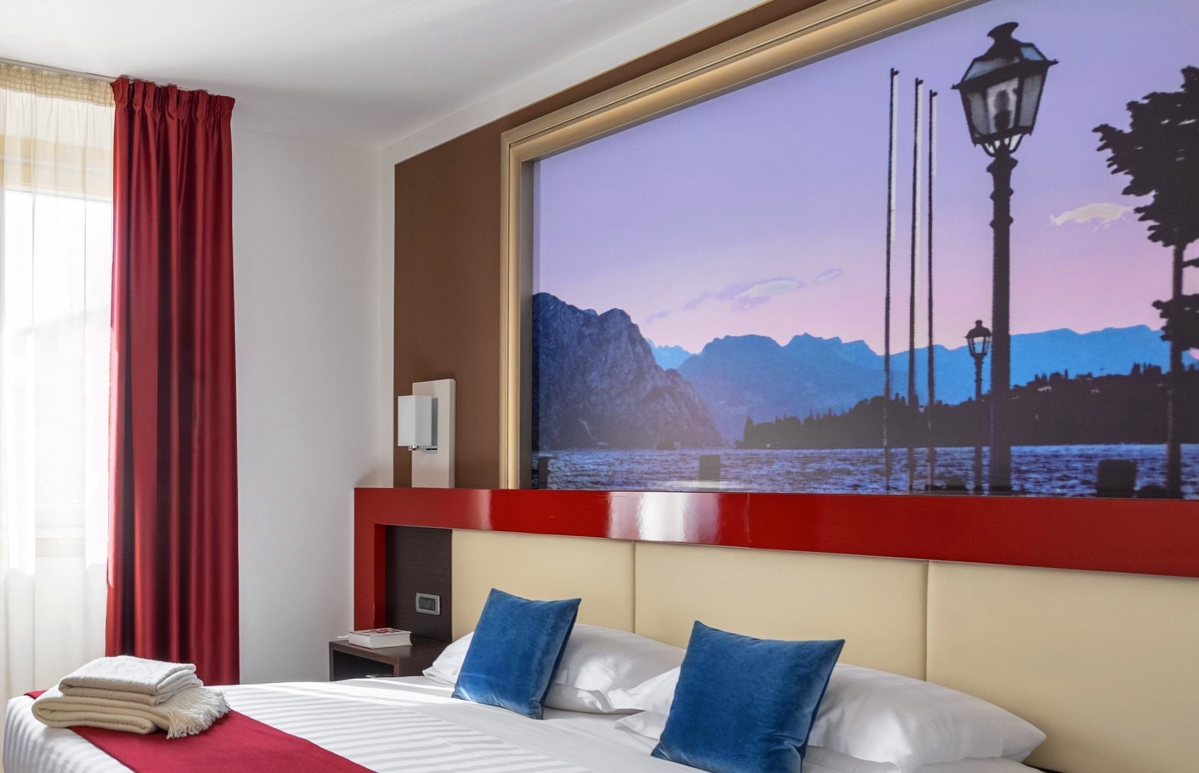 A hotel room with a bed, pillows, a nightstand, a TV mounted on the wall, and a window with curtains.