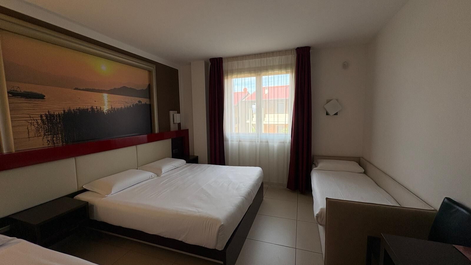 A hotel room with a bed, a desk, a chair, a painting on the wall, and a window with curtains.