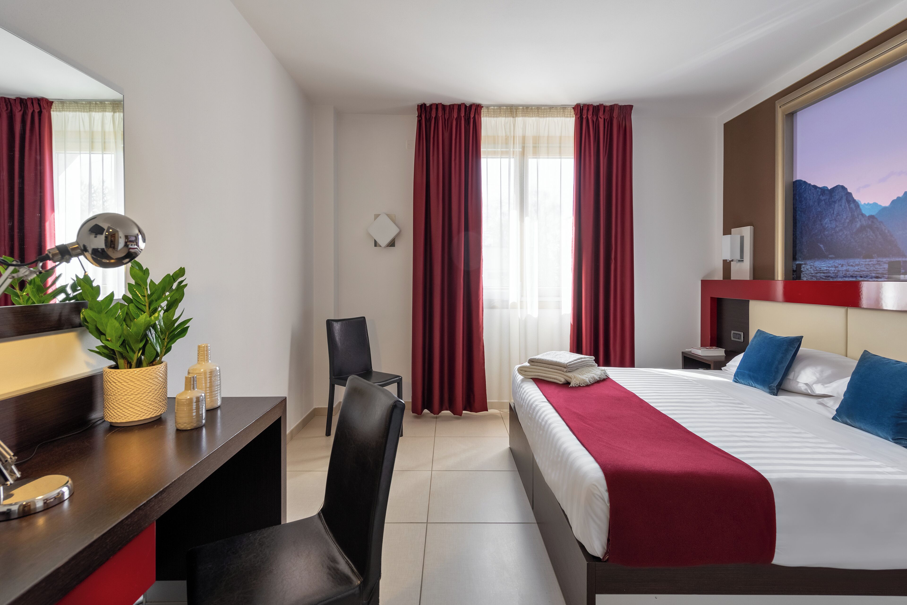 A hotel room with a bed, a desk with a mirror, a chair, a plant, and a TV on the wall.