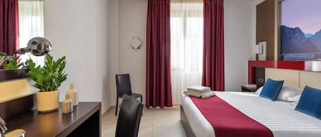 A hotel room with a bed, a desk with a mirror, a chair, a plant, and a TV on the wall.