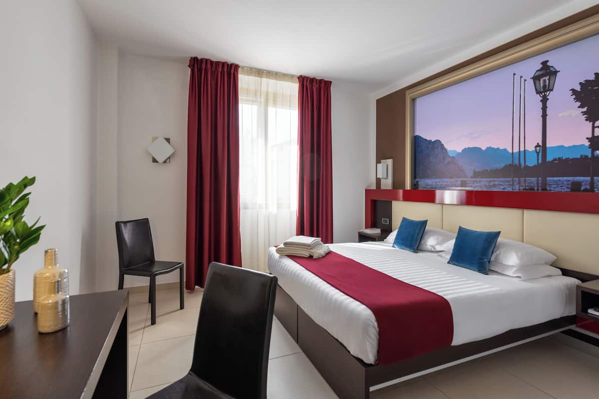 A hotel room with a bed, a desk, a chair, a table, a lamp, and a painting on the wall.