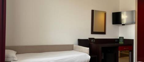Habitación de hotel con una cama individual, un escritorio con una silla, un televisor de pantalla plana montado en la pared y una mesita con un libro.