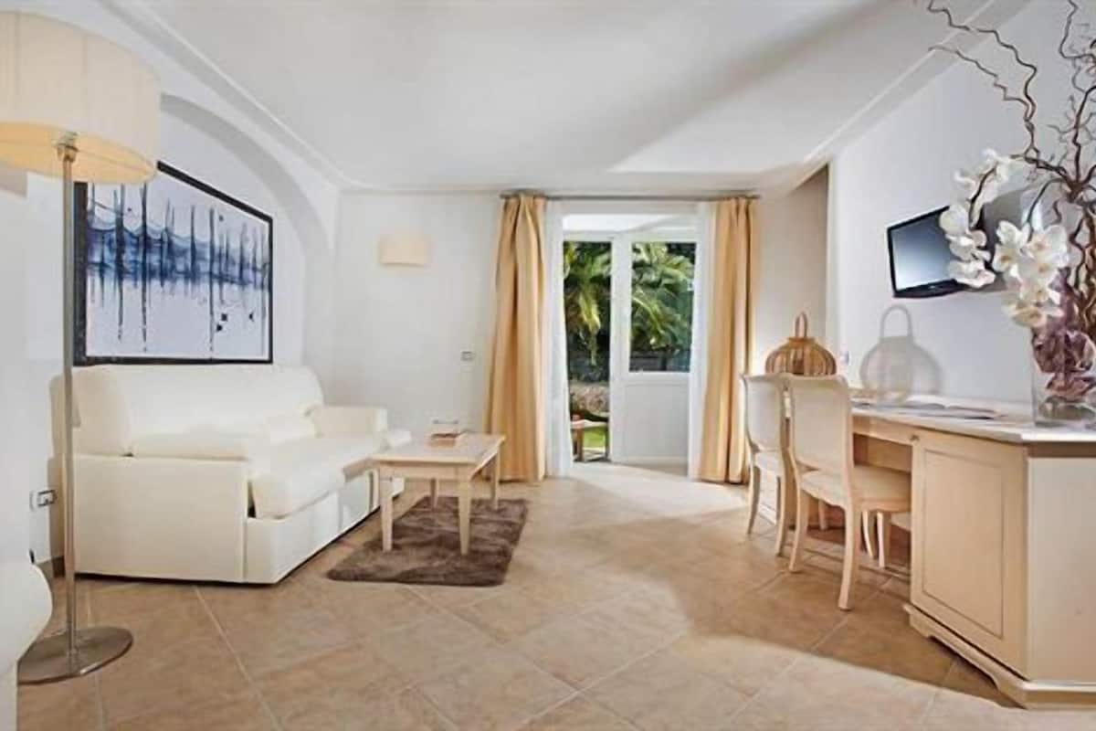 A modern living room with a white sofa, a small table, a desk with a chair, a TV, and a large window with curtains.